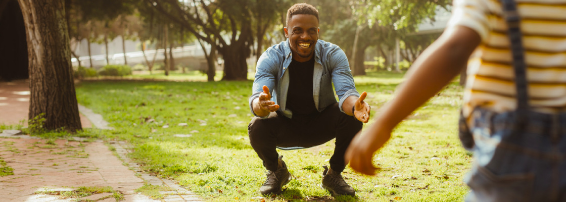 A father squatting down at the park with his arms open embracing his son running to him
