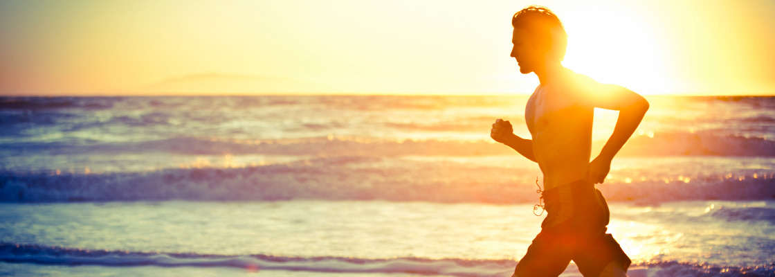 Side profile view of man jogging along the beach with the ocean and bright sun shine behind him