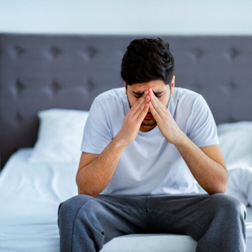 Younger man sitting at the bed side, with his hands clasped over his head, enduring pain