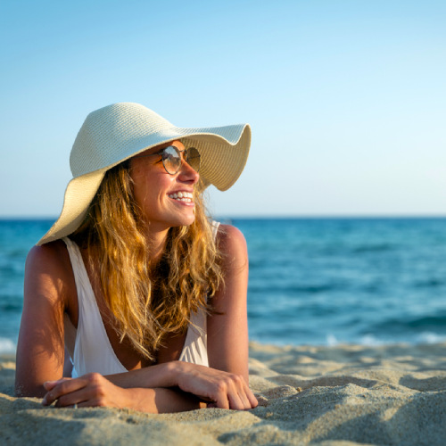 A mature woman with glasses wearing a hat is seen laying on the beach at sunset, happily smiling